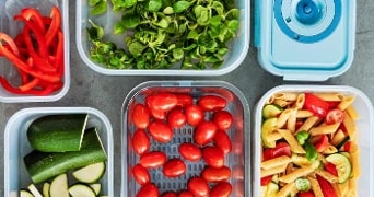 Vers gesneden paprika, veldsla, courgette, cherrytomaten en pastasalade in plastic opbergdozen.