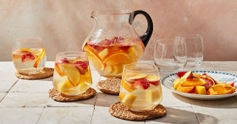 Kan met fruit belegde pitcher en drie glazen met frisdrank tonen, op een tafel gedecoreerd met een schaal fruit.