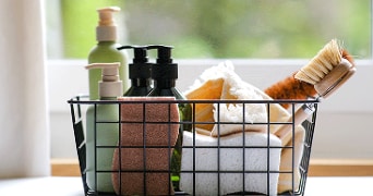 Mand draadmandje met schoonmaakartikelen zoals handzeepdispensers, sponsen, borstels en handdoeken, geplaatst voor een raam.