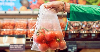 Herbruikbaar groentenzakje met verse tomaten erin in de hand van een vrouw met een groene jas en op de achtergrond de versafdeling van een supermarkt