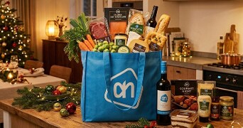Albert Heijn boodschappentas gevuld met diverse feestelijke producten zoals groenten, vlees, stokbrood, kaas, wijn en gourmet artikelen op een keukentafel, met kerstdecoratie op de achtergrond.