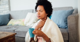 Persoon met zwarte krullen drinkt uit een blauwe mok zittend op een bank met een deken omgeslagen.
