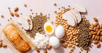 Zalmfilet, kipfilet, gekookte eieren, cottage cheese, linzen, kikkererwten en amandelen op een lichtroze ondergrond, als voorbeeld van eiwitrijke voedingsmiddelen.