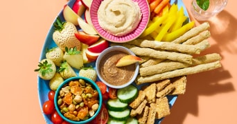 Gezond borrelplateau met diverse groenten, fruit, crackertjes, groentesticks, hummus, notendip en snackgroenten, gepresenteerd op een blauw bord.