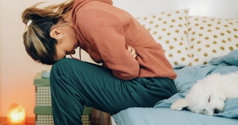 Persoon in een roodbruine trui zit voorovergebogen op bed naast een slapende witte hond.