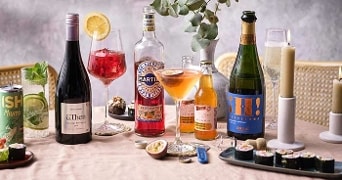 Assortiment van verschillende alcoholische dranken en cocktails op een gedekte tafel, inclusief een Mojito blikje van ISH, een fles El Then wijn, Martini Vibrante aperitief en een fles Hi! Cava, omringd door sushi en kaarsen voor een feestelijke setting.