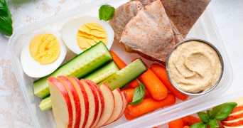 Lunchbox met gesneden appel, komkommer, wortels, gekookt ei, volkoren pita, en een bakje hummus.