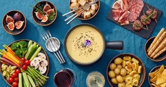 Gourmet kaasfondue op een blauwe tafel, omringd door groenten, brood, aardappelen, tortellini, dipstokjes, vijgen, vleeswaren en twee glazen wijn.