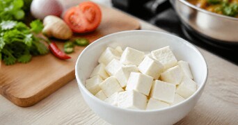 Blokjes tofu in een witte kom met verse groenten, waaronder tomaat, koriander, peper en ui, op een houten snijplank op de achtergrond.