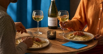 Twee personen genieten van een diner met witte asperges, ham en witte wijn aan een ronde tafel, met een fles Sancerre wijn en een brandend kaarsje op de achtergrond.