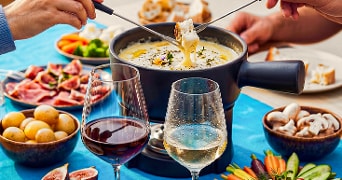 Fondue met kaas in fonduepan, omringd door gesneden groenten, aardappelen, brood, vleeswaren en glazen rode en witte wijn.