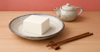 Blok tofu op een bord met houten eetstokjes en een keramische theepot op een houten tafel.