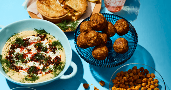 Assortiment van vegetarische borrelhapjes, waaronder hummus met kruiden en olie, falafelballetjes, geroosterde kikkererwten en een hartige taart, geserveerd op een blauwe ondergrond.