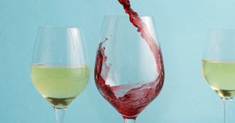 Twee glazen witte wijn en een glas waar rode wijn in wordt geschonken, tegen een blauwe achtergrond.