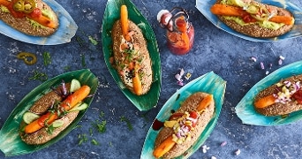 Geroosterde wortelhotdogs met diverse toppings zoals ketchup, mosterd, augurk en rode ui, geserveerd in bladerdeeg op een blauw bord.