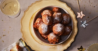 Kom met oliebollen bestrooid met poedersuiker op een feestelijke tafel, omringd door glazen met champagne en feestelijke decoraties.