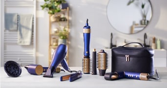 Set haarstyling tools in blauw en goud, waaronder een föhn, opzetborstels, krultang, stijltang en een zwarte opbergcase, uitgestald op een tafel in een moderne badkamer.
