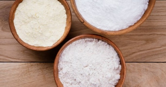 Drie houten kommen gevuld met verschillende poeders, waaronder bloem en suiker, op een houten tafel.