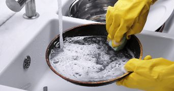 Afwassen van een koekenpan met schuimend sop in de wasbak, gedragen door gele rubberen handschoenen.