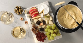 Bord met diverse kazen, stukjes brood, appel, peer, druiven, walnoten en olijven, naast een pot kaasfondue en twee glazen witte wijn.