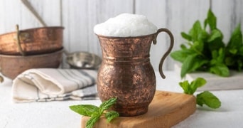 Koperen beker gevuld met schuimige ayran, met een takje munt ernaast op een houten snijplank. Achtergrond met metalen schalen en verse muntbladeren.