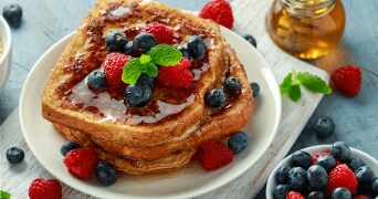 Gouden wentelteefjes met blauwe bessen, frambozen en munt, overgoten met siroop op een wit bord