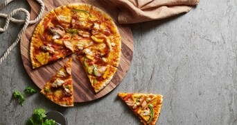 Houten serveerplank met barbecuekip pizza en een losse pizzapunt op een beige tafel.