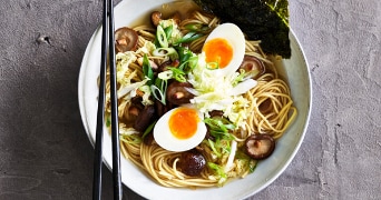 Kom ramen met noedels, perfect gekookt ei, shiitake-paddenstoelen, verse groenten en nori, geserveerd met zwarte eetstokjes.