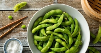 Kom met verse edamame bonen bestrooid met zoutvlokken op een houten tafel, met eetstokjes en een kom zout.