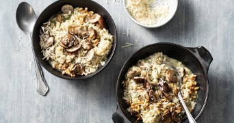 Risotto met gebakken champignons, pijnboompitten en geraspte kaas geserveerd in kommen op een grijze ondergrond