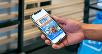Hand met smartphone waarop een Bonuskaart van Albert Heijn wordt weergegeven met barcode en afbeeldingen van fruit en groenten.