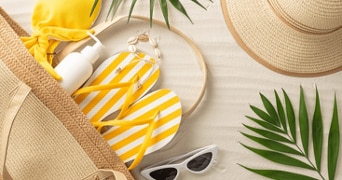 Zomerse strandbenodigdheden met een strooien tas, gele gestreepte slippers, zonnebrandcrème, zonnebril en een strooien hoed op zand met groene bladeren.