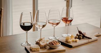 Vier glazen wijn, variërend van rood tot rosé, geserveerd met kaas, brood, olijven, en dip op houten planken.