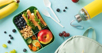 Lunchbox met broodjes, appel, blauwe bessen, cashewnoten, komkommer, wortel en een fles water, met bananen en druiven op een lichtblauwe achtergrond naast een rugzak