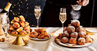 Feestelijke tafel met oliebollen, beignets, champagneglazen en lichtjes op de achtergrond.