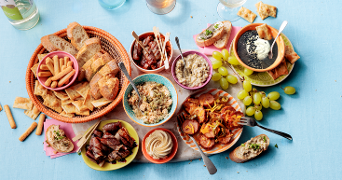Tapasplank met verschillende soorten brood, dips, gegrilde groenten, olijven, druiven en worst op een lichtblauwe ondergrond.