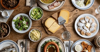 Typisch Nederlandse maaltijd met verschillende gerechten op een houten tafel, waaronder kaas op een kaasplank, stamppot met rookworst, salade, poffertjes met poedersuiker en boter, gerookte haring, vleeswaren, aardappelpuree en een bordje kibbeling met saus.
