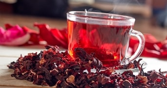 Kopje hibiscusthee omringd door gedroogde hibiscusbloemen op een houten tafel.