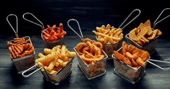 Verschillende soorten friet, zoals krulfriet, aardappelschijfjes en zoete aardappelfriet, geserveerd in metalen mandjes op een donkere houten tafel.