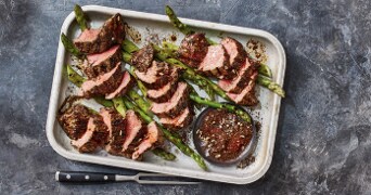 Gesneden gegrild vlees op een schaal met groene asperges en een kommetje kruiden.
