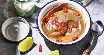 Kom pindasaus gegarneerd met rode chilipepers, naast een potje saus, limoenschijfjes en een lepel.