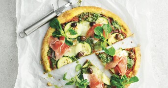Pizza met parmaham, courgette, olijven en veldsla op bakpapier, met een pizzasnijder.