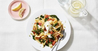 Een bord met pappardellepasta, spinazie, tomaten, roomkaas en pistachenoten naast een roze bordje met citroenpartjes en een karaf water met schijfjes citroen.