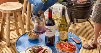 Tafel met flessen rode en witte wijn (waaronder Casillero del Diablo), glazen wijn, een bord met vlees, een hotdog met groenten, een bakje olijven en een barbecue op de achtergrond.
