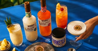 Cocktails en dranken op een blauwe tafel, waaronder twee flessen van de merken KUPFERBERG voor Piña Colada en Sex on the Beach cocktail, een Pornstar Martini pot, een glas Piña Colada gegarneerd met ananas, een glas Sex on the Beach cocktail met sinaasappel en kers, en een glas Pornstar Martini met halve passievrucht.