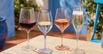 Vier wijnglazen op een houten tafel met rode wijn, witte wijn, rosé en mousserende wijn in een zonnige buitenomgeving.