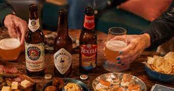 Verschillende flesjes speciaalbier van de merken Paulaner, Brouwerij ’t IJ en Texels op een houten tafel, omringd door borrelhapjes, kaas, chips en mensen die bier inschenken.