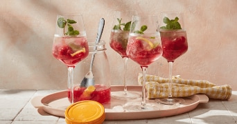 Vier glazen met roze cocktail, geserveerd met citroen, munt en ijs op een dienblad, naast een glazen pot met fruitmix en een gele doek.