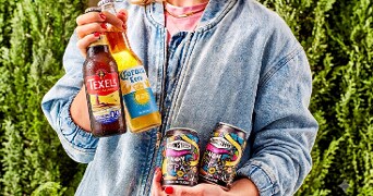 Vrouw in spijkerjasje houdt alcoholvrije bieren vast, waaronder Texels 0.0%, Corona Cero en twee blikken Vandestreek Non Alcoholic IPA, met groene struiken op de achtergrond.