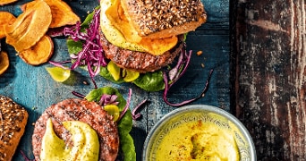 Twee sappige hamburgers met sla, rode kool, avocado, zoete aardappelchips en gele saus op een donkerhouten tafel, met een kommetje saus erbij.